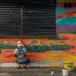 Artista pinta um mural colorido ao ar livre, em uma cena de produção cultural.