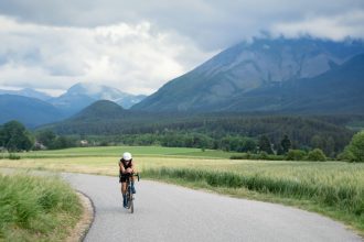 Ciclista percorre uma estrada rural sinuosa, cercada por vegetação e montanhas ao fundo.