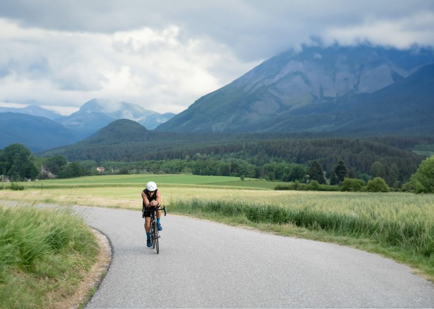 Ciclista percorre uma estrada rural sinuosa, cercada por vegetação e montanhas ao fundo.