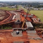 Vista aérea de uma obra de construção rodoviária, com máquinas trabalhando em meio à paisagem.